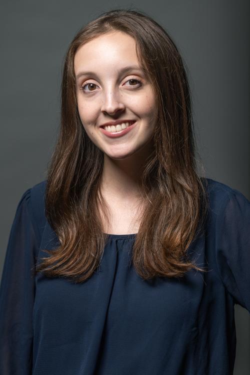 Smiling young woman with long brown hair in a dark blue top, gray background.